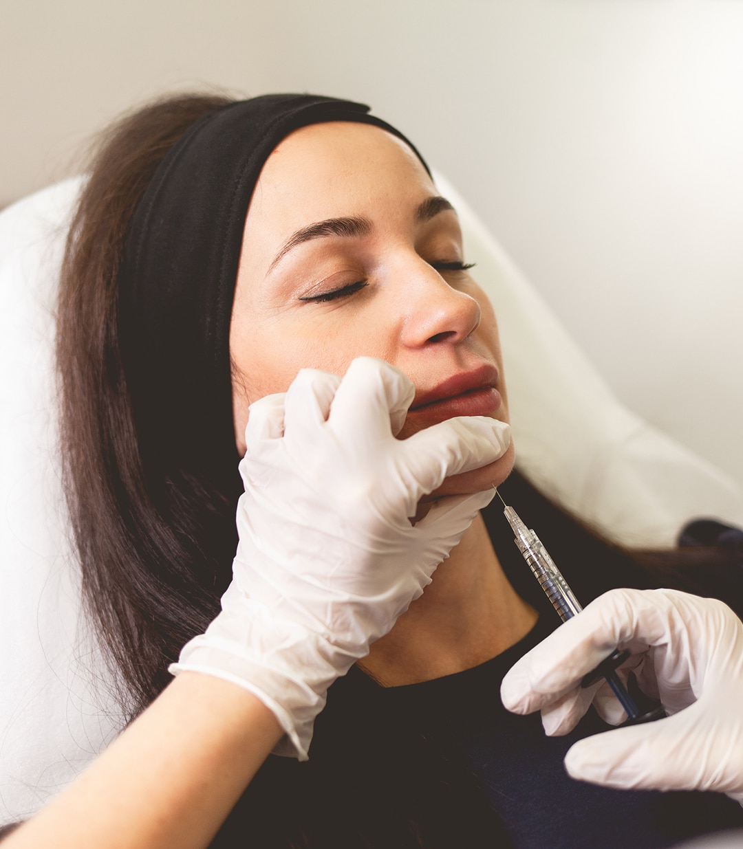 A woman receiving a Kybella injection in the chin area while lying down, performed by a medical professional wearing white gloves.