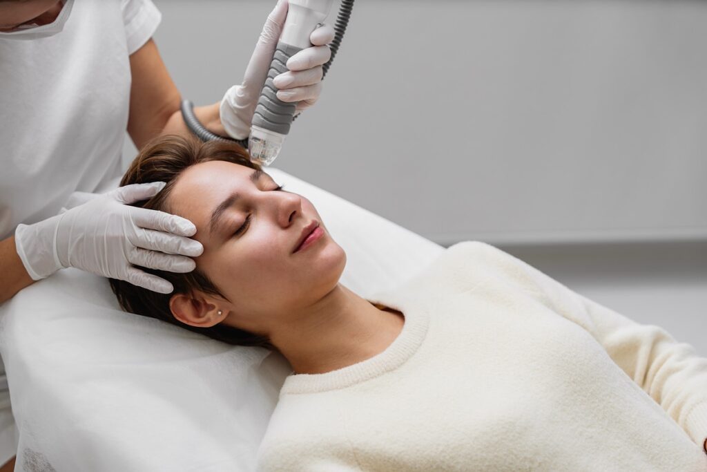 A woman is lying on a treatment bed with her eyes closed while a skincare professional performs an RF microneedling procedure on her forehead using a handheld device.