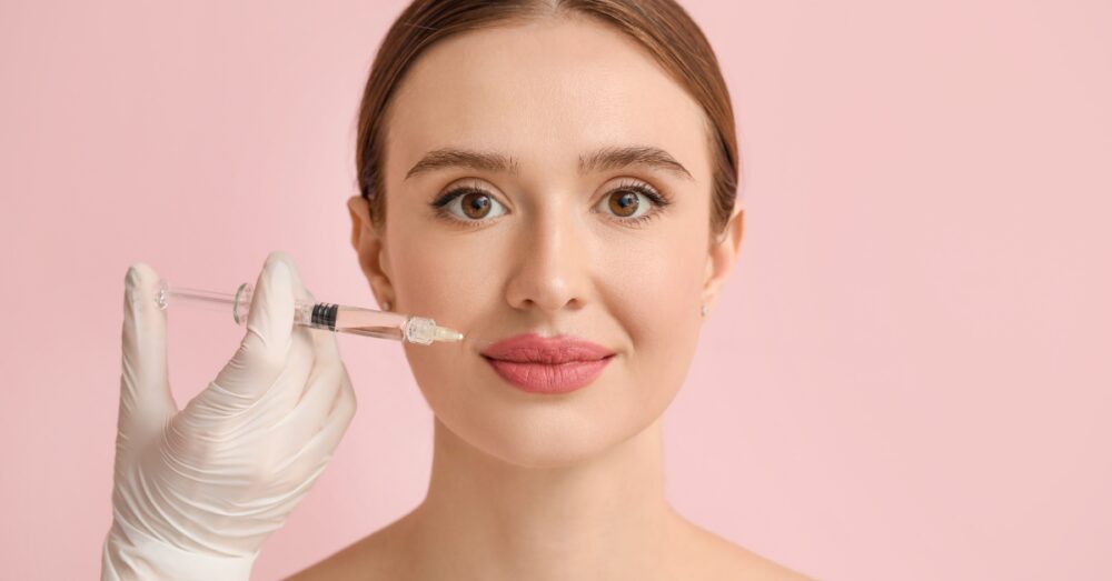 A young woman with brown hair pulled back smiles softly as a gloved hand holds a syringe near her upper lip area against a pink background
