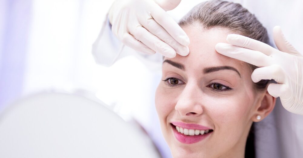 Woman looking into a mirror while smiling as a gloved provider touches her forehead